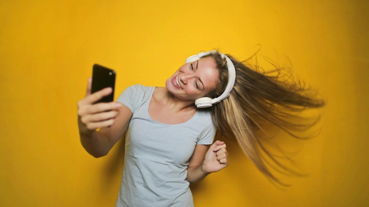 Cheerful woman wearing headphones, enjoying music and taking a selfie against a vibrant yellow background. via Pexels