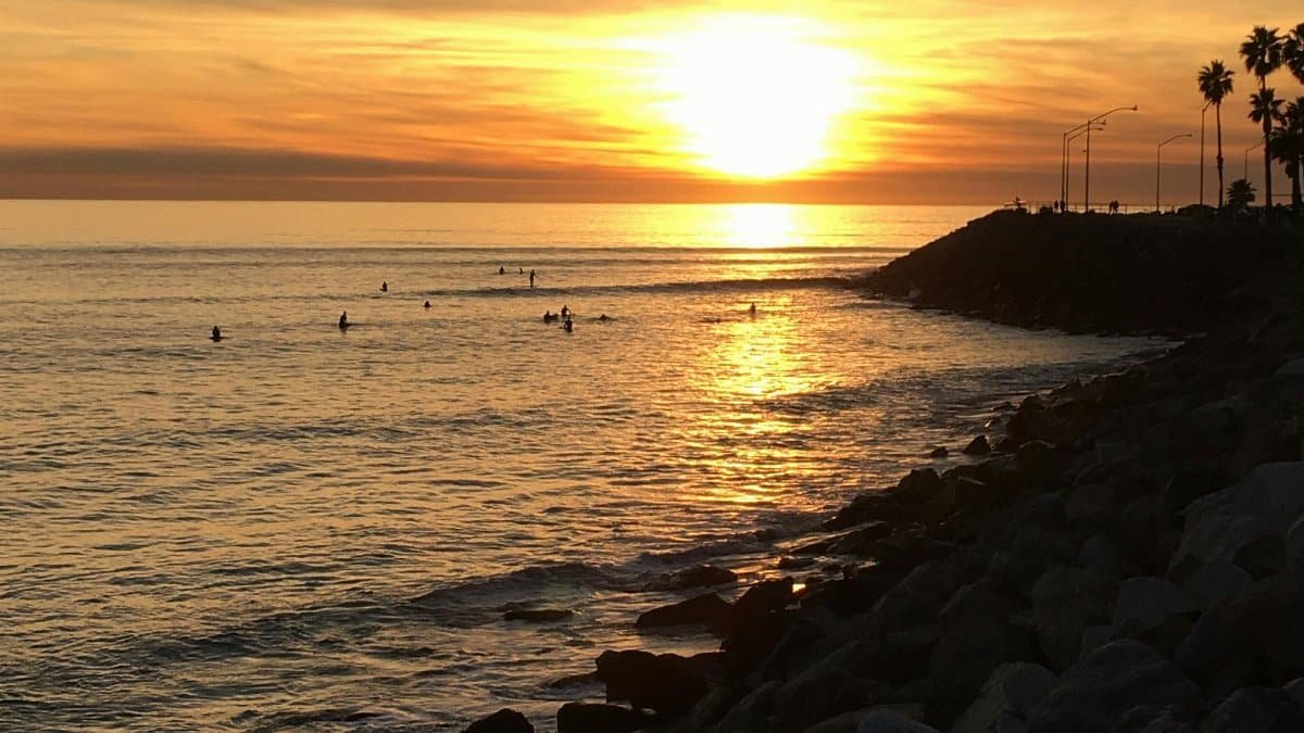 A stunning sunset over the Pacific Ocean with surfers enjoying the waves near Santa Monica's rocky coastline.