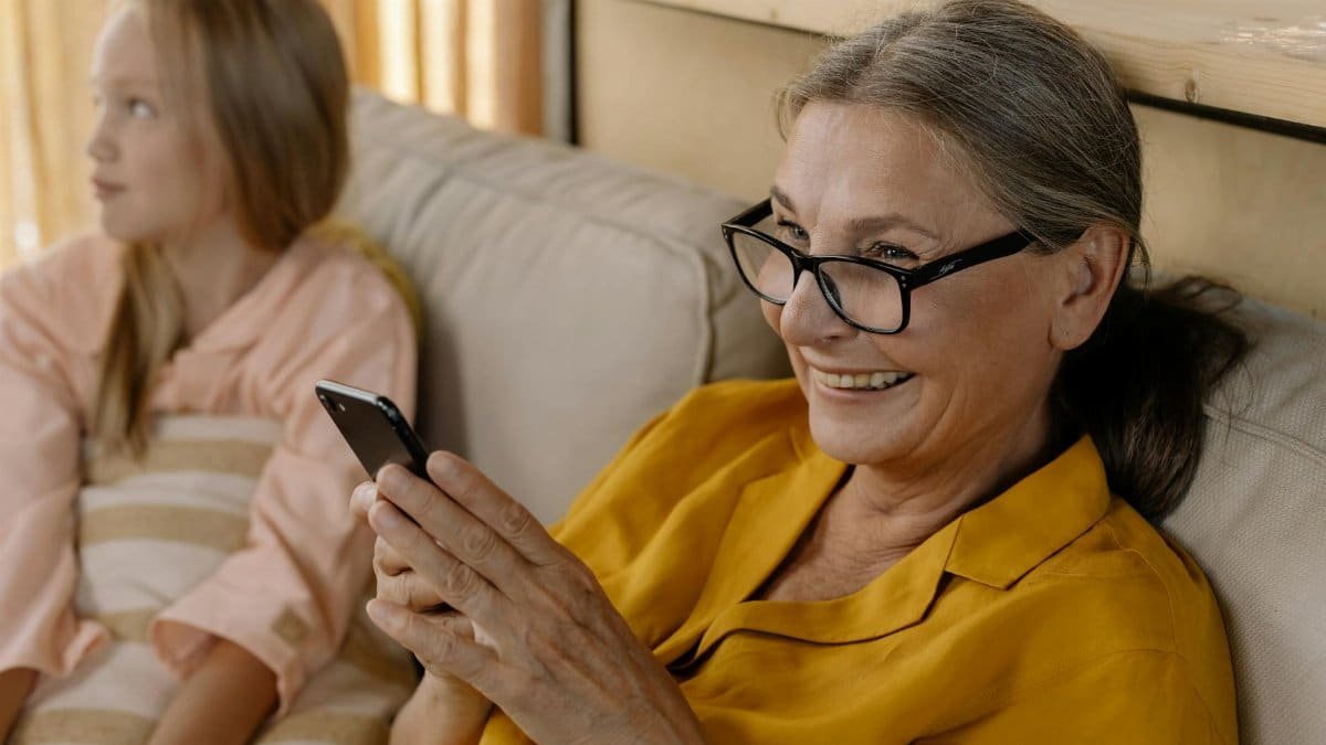 Senior woman in glasses smiling while using smartphone indoors, sitting with a child.