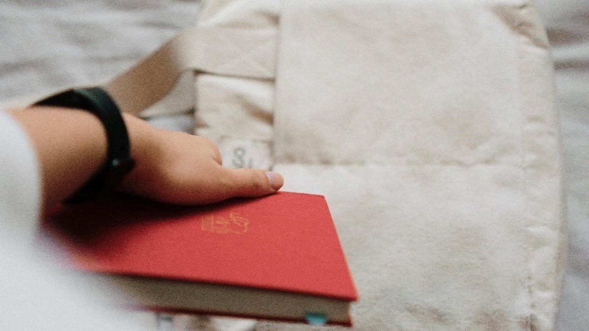 A close-up of a hand wearing a watch, holding a red book on a soft beige surface.