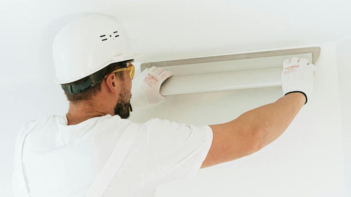 A construction worker wearing safety gear installs a light fixture in a modern indoor setting.