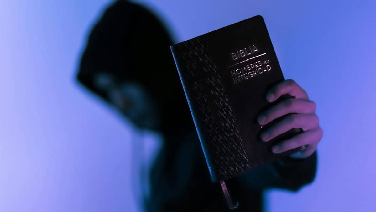 A hooded figure holds a Bible in low-light setting, emphasizing spirituality and mystery.