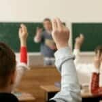 Students in a classroom raising hands, engaging in a lesson with the teacher.