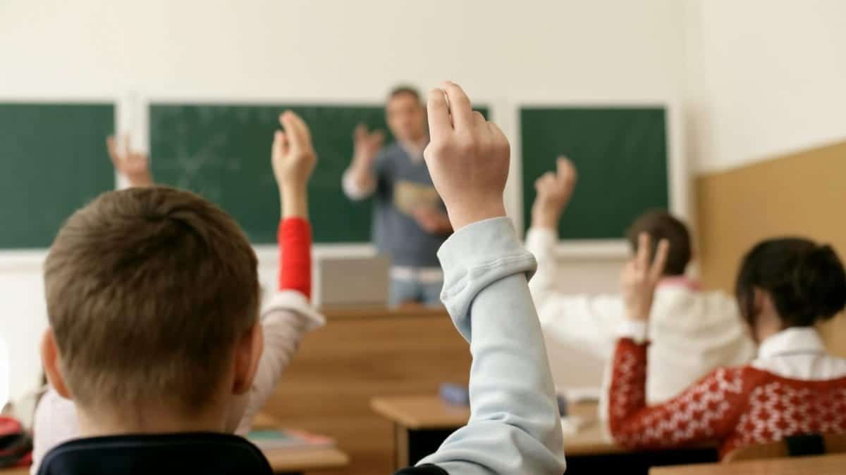 Students in a classroom raising hands, engaging in a lesson with the teacher.