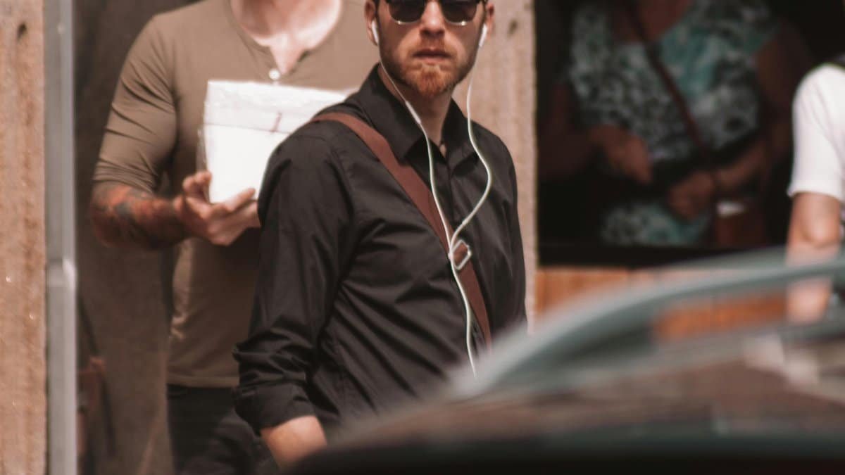 A stylish man in black long sleeves and sunglasses with earphones stands by a car on a sunny street. via Pexels