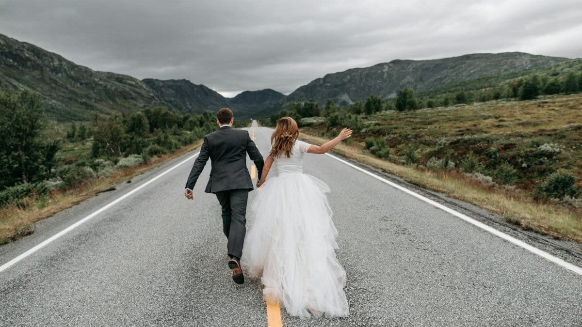 Bride and groom holding hands, running along a scenic mountain road. Capturing love and adventure.