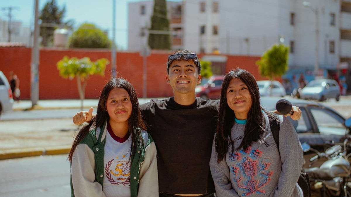 Three smiling friends posing together in an urban outdoor setting on a sunny day.