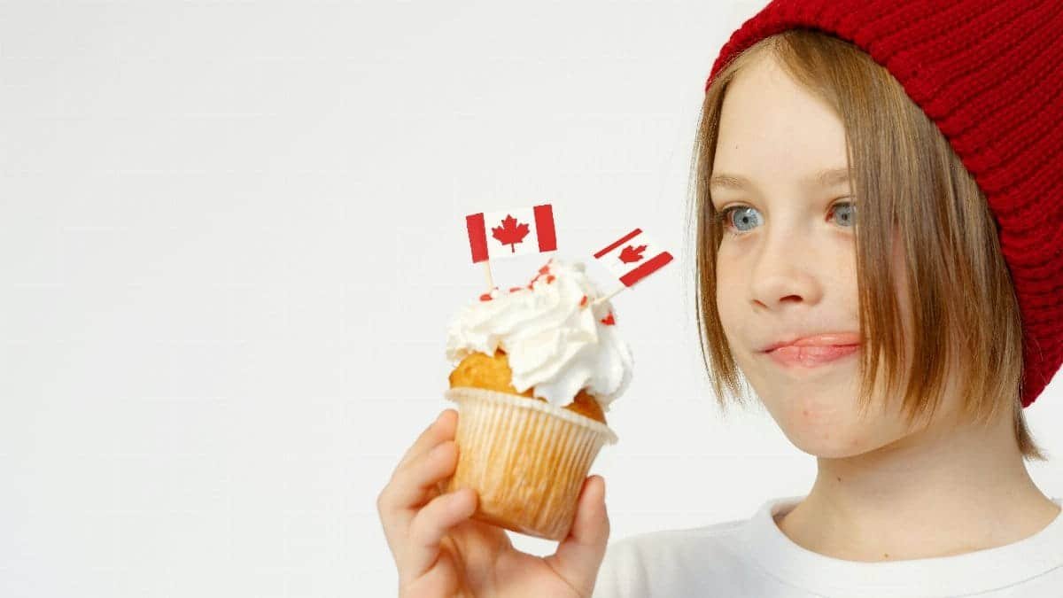 A child wearing a red beanie, holding a cupcake topped with Canadian flags and whipped cream.