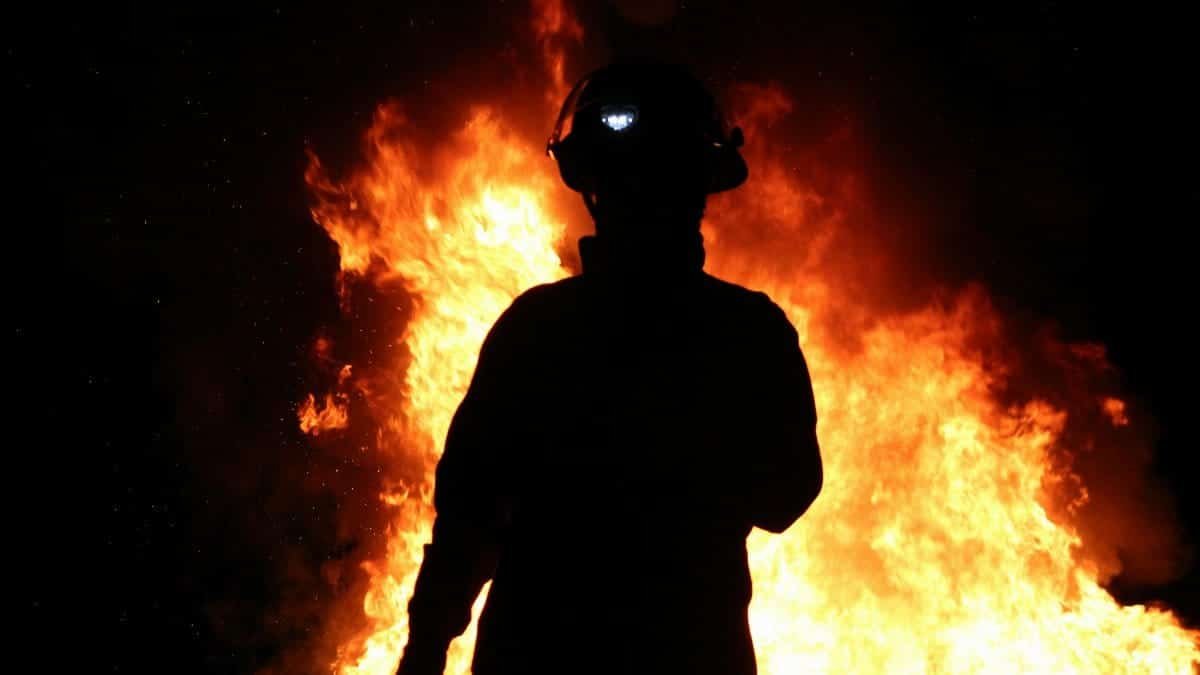 A silhouetted firefighter stands against a backdrop of roaring flames at night.