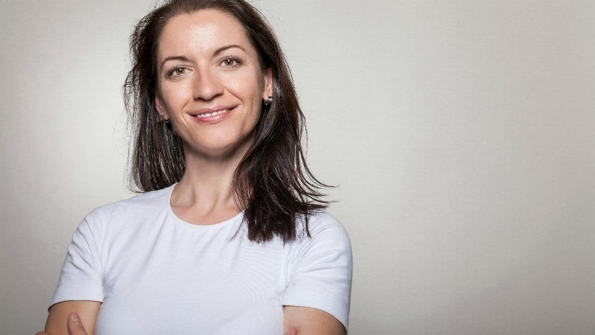 Portrait of a confident woman smiling with arms crossed, wearing a white t-shirt on a gray background.