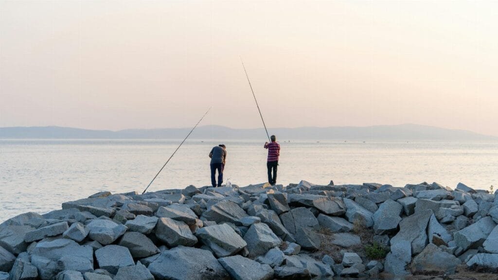 Two men fishing on a rocky shoreline as the sun rises, casting long shadows over a tranquil sea.