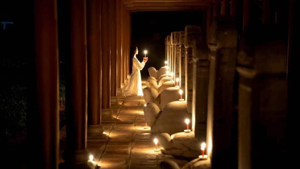 Side view of unrecognizable female with long dark hair in white dress standing on knees with burning candle near statue in temple in darkness
