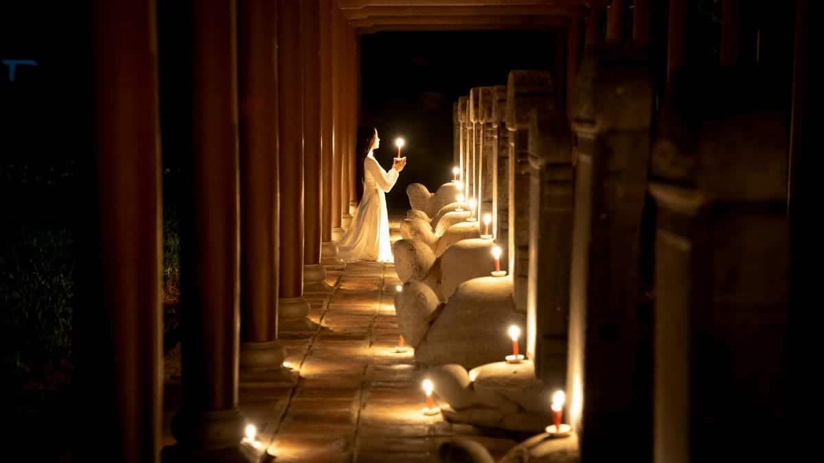 Side view of unrecognizable female with long dark hair in white dress standing on knees with burning candle near statue in temple in darkness