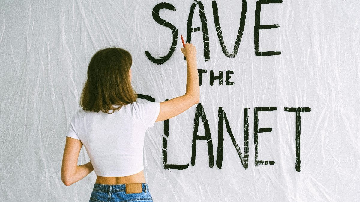 A woman in jeans and white top paints 'Save the Planet' on a wall, advocating environmental awareness. via Pexels