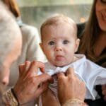 A touching moment of an Orthodox baptism ceremony featuring a smiling baby and a priest.