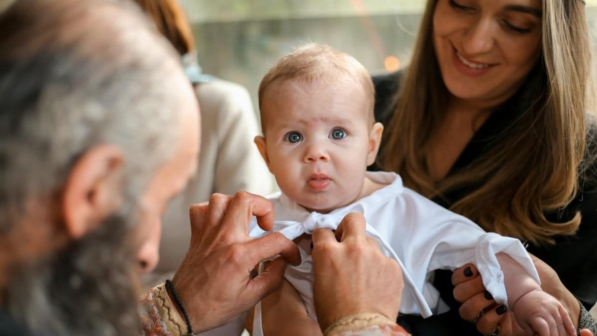 A touching moment of an Orthodox baptism ceremony featuring a smiling baby and a priest.