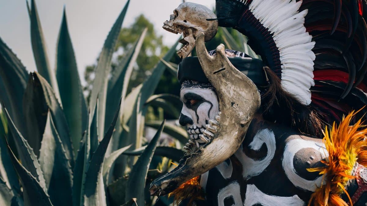 A vividly dressed man in Day of the Dead costume with skull headdress, outdoors in Mexico.