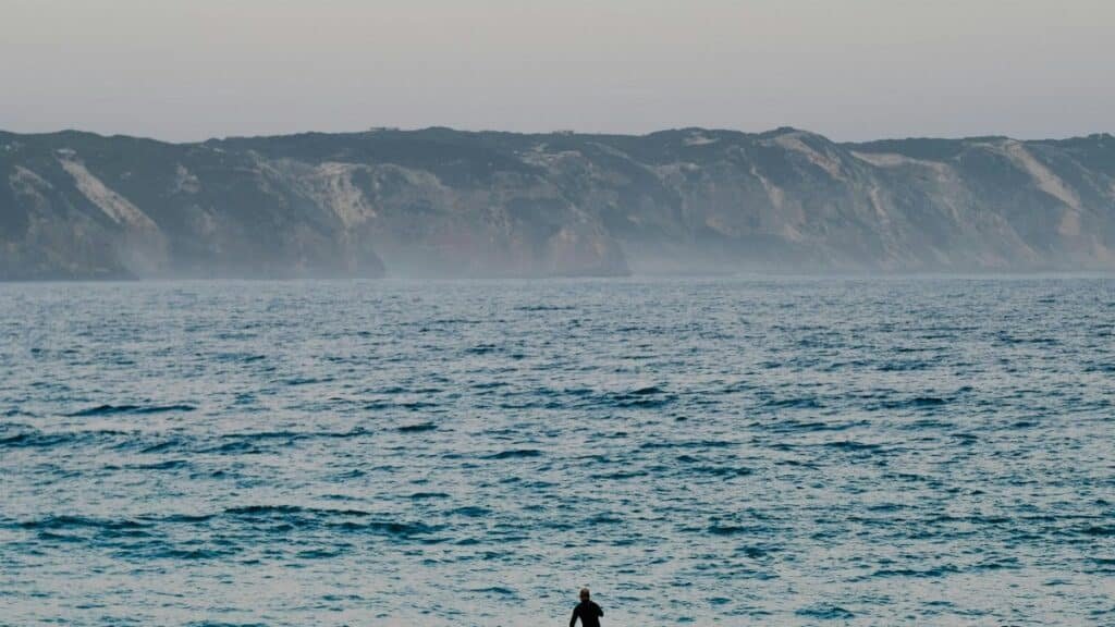 A lone paddleboarder navigates calm waters at sunset, with distant cliffs under a soft sky. via Pexels