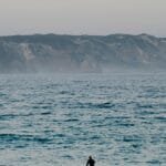A lone paddleboarder navigates calm waters at sunset, with distant cliffs under a soft sky. via Pexels