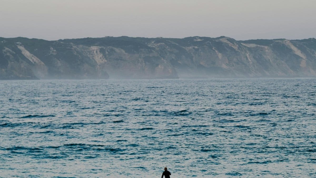 A lone paddleboarder navigates calm waters at sunset, with distant cliffs under a soft sky. via Pexels