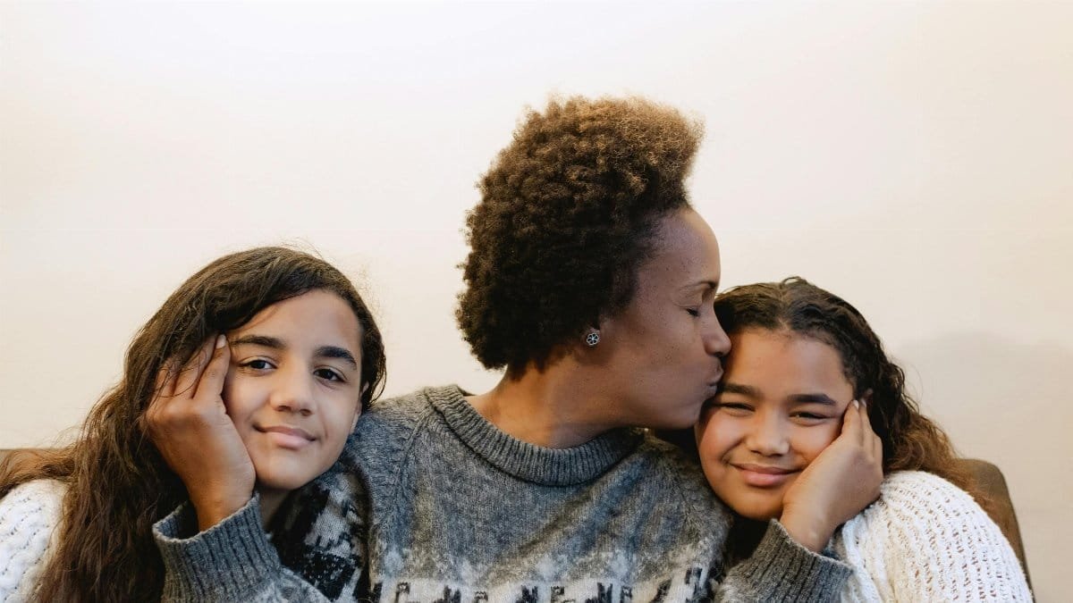 A joyful mother kissing her smiling daughters indoors, capturing love and family warmth. via Pexels