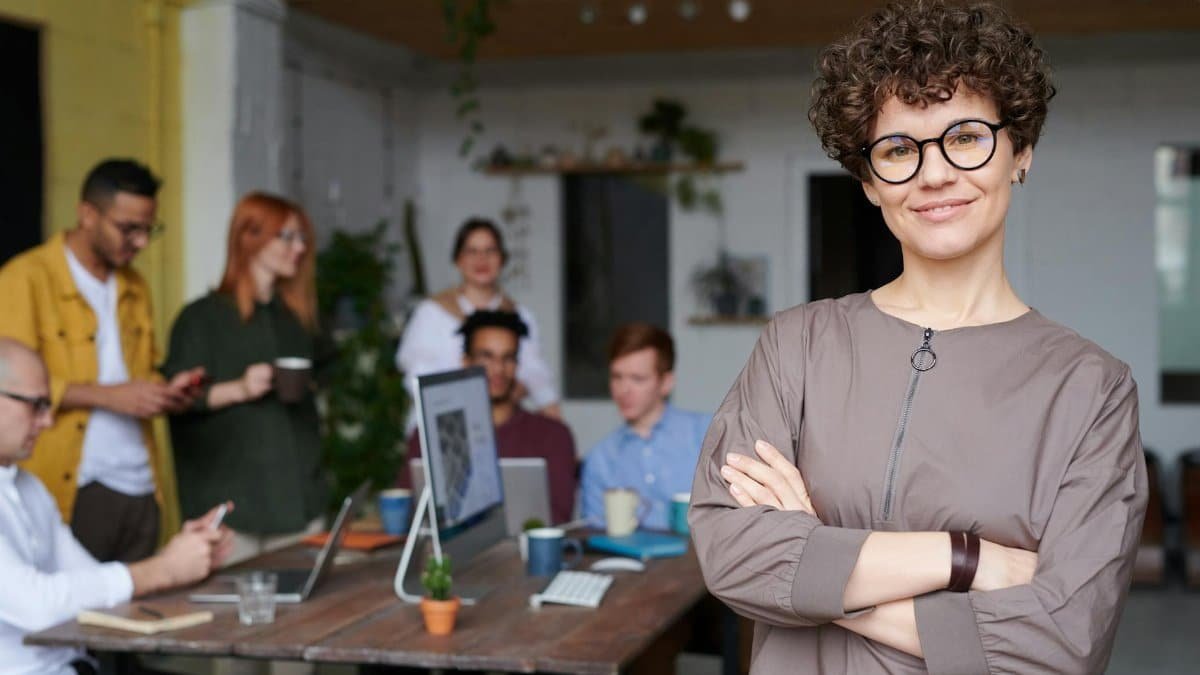 Smiling businesswoman with curly hair stands confidently in a modern office space with colleagues.