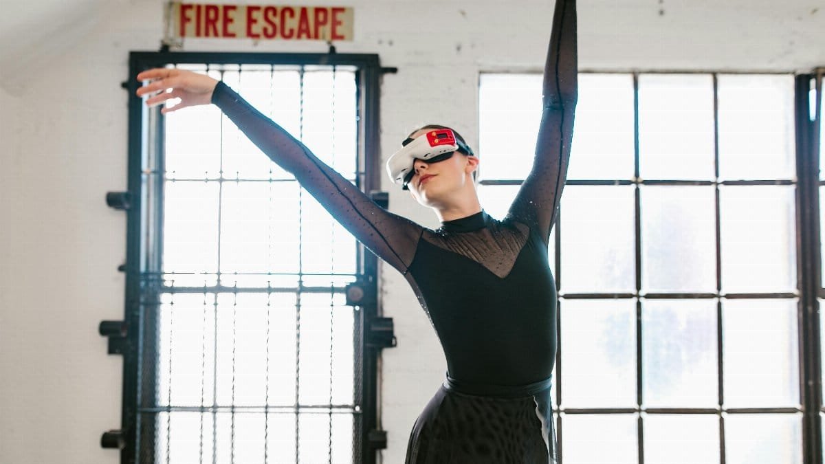 A female ballet dancer in a black outfit using a VR headset, striking a graceful pose indoors. via Pexels