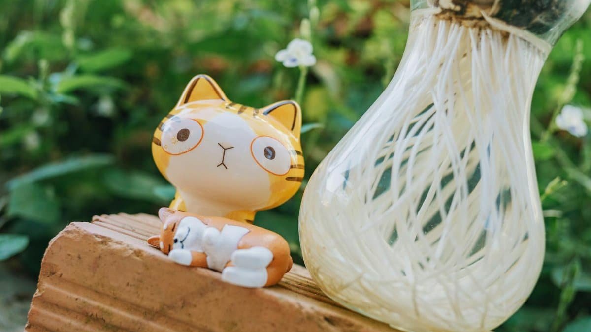 Charming ceramic cat figurine next to a glass vase with roots, set in a lush outdoor environment.