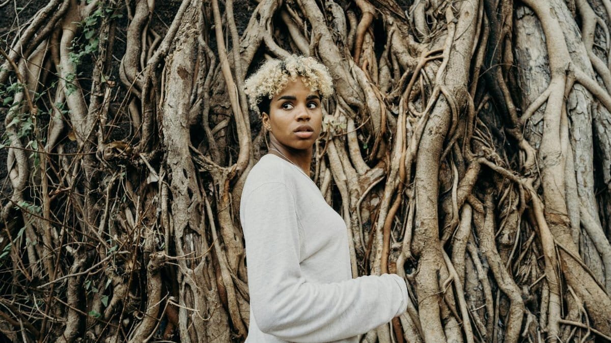 A young woman with curly hair stands in front of intricately twisted tree roots, showcasing a natural backdrop.