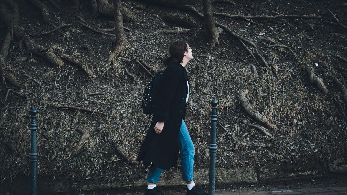 A woman in a long coat walks on a sidewalk surrounded by exposed tree roots.