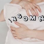 Close-up of a woman holding a pillow with 'insomnia' text, depicting sleep disorder. via Pexels