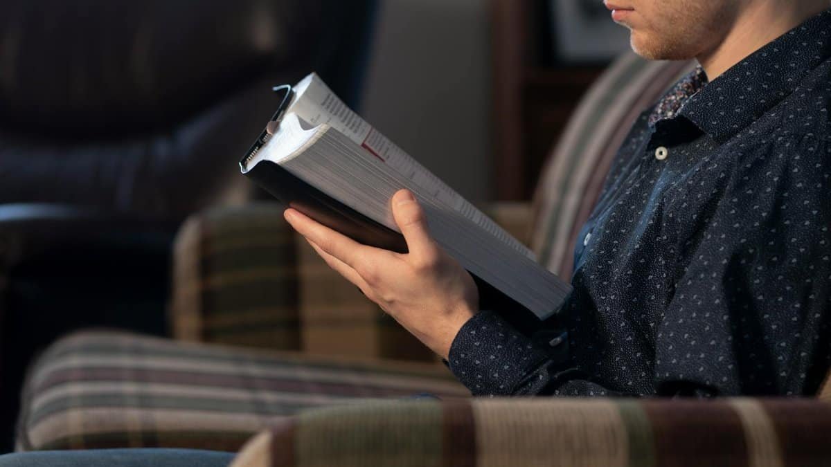 Adult male reading a Bible while seated on a comfortable chair indoors.