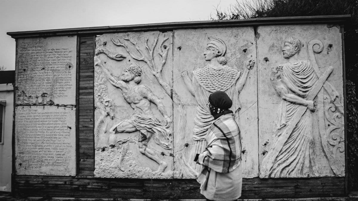 A woman walks past detailed ancient stone relief sculptures outdoors, captured in black and white.