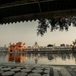 Sidewalk with roof near water with illuminated ancient buildings with fence against gray sky in city street in evening time