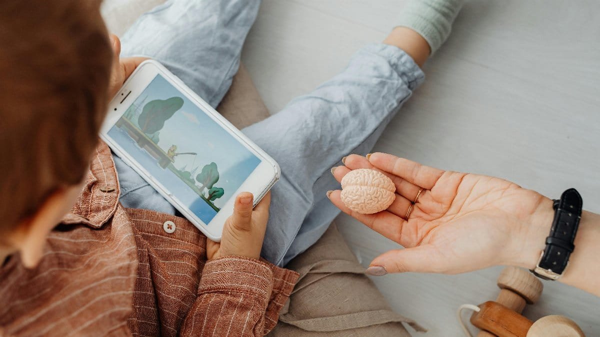 Young child engrossed in a smartphone video while holding a brain model indoors.