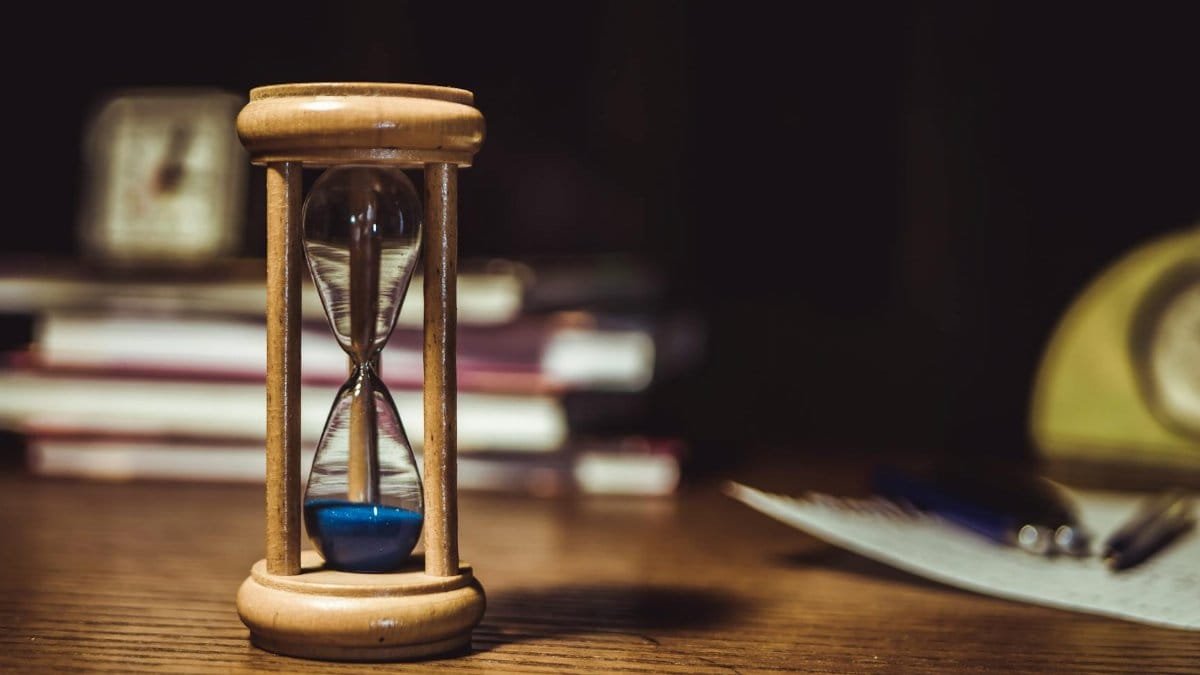 A close-up of a wooden hourglass with blue sand on a wooden desk, symbolizing time and patience.