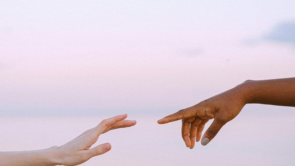 Two diverse hands reaching towards each other over a serene pastel sky and water background. via Pexels