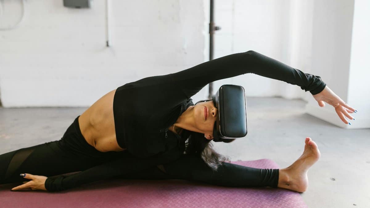 Woman in activewear using VR goggles for yoga stretching on a mat indoors.