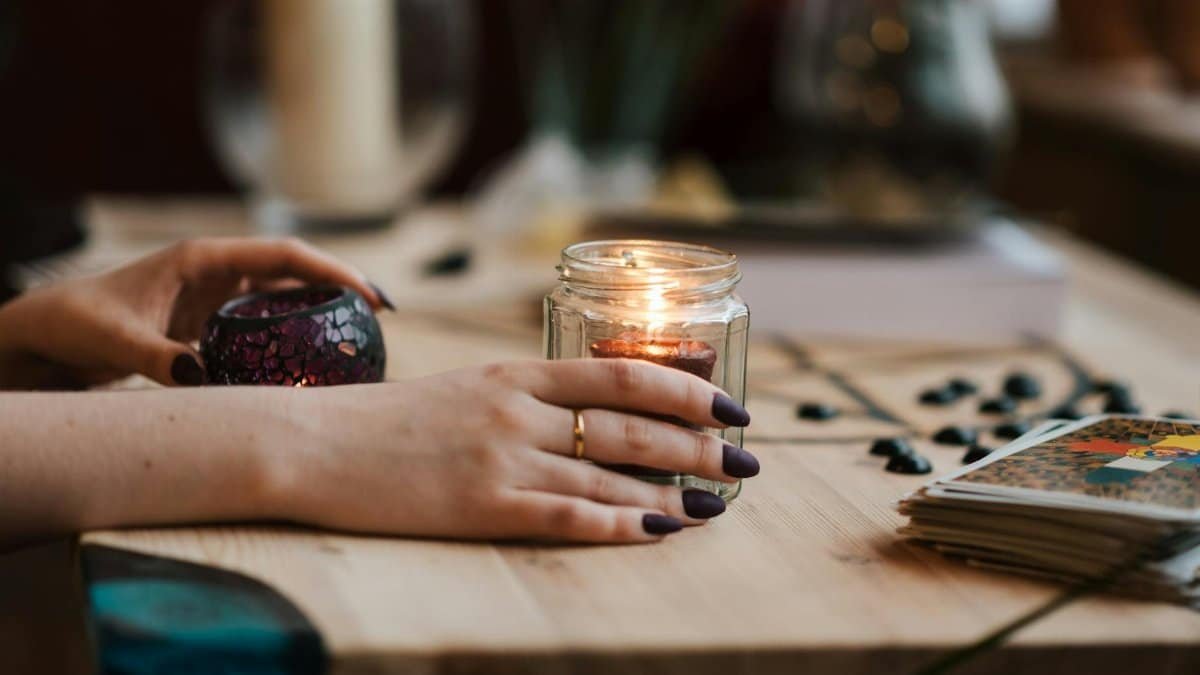 Hands with dark nails holding a lit candle, surrounded by tarot cards, creating a mystical and warm ambiance.