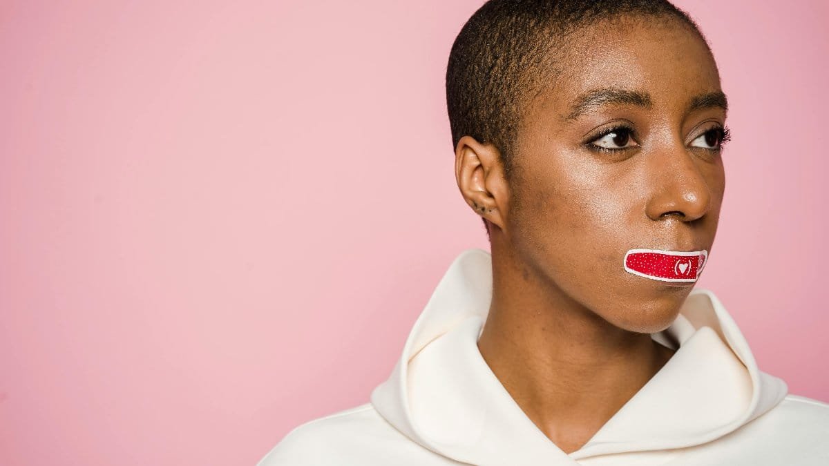 Empowered African American woman with Band-Aid on lips, symbolizing silence and awareness on a pink background.