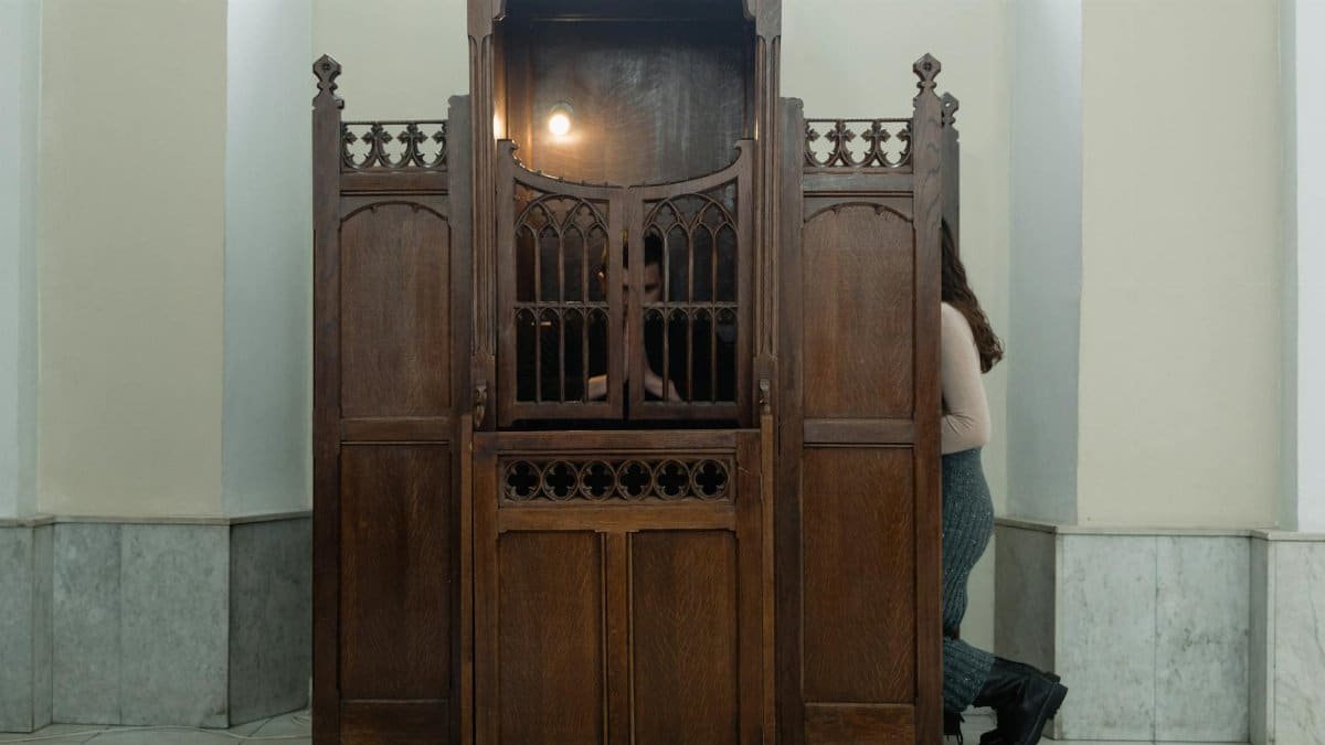 A wooden confessional booth in a church with a person sitting inside, partially visible.