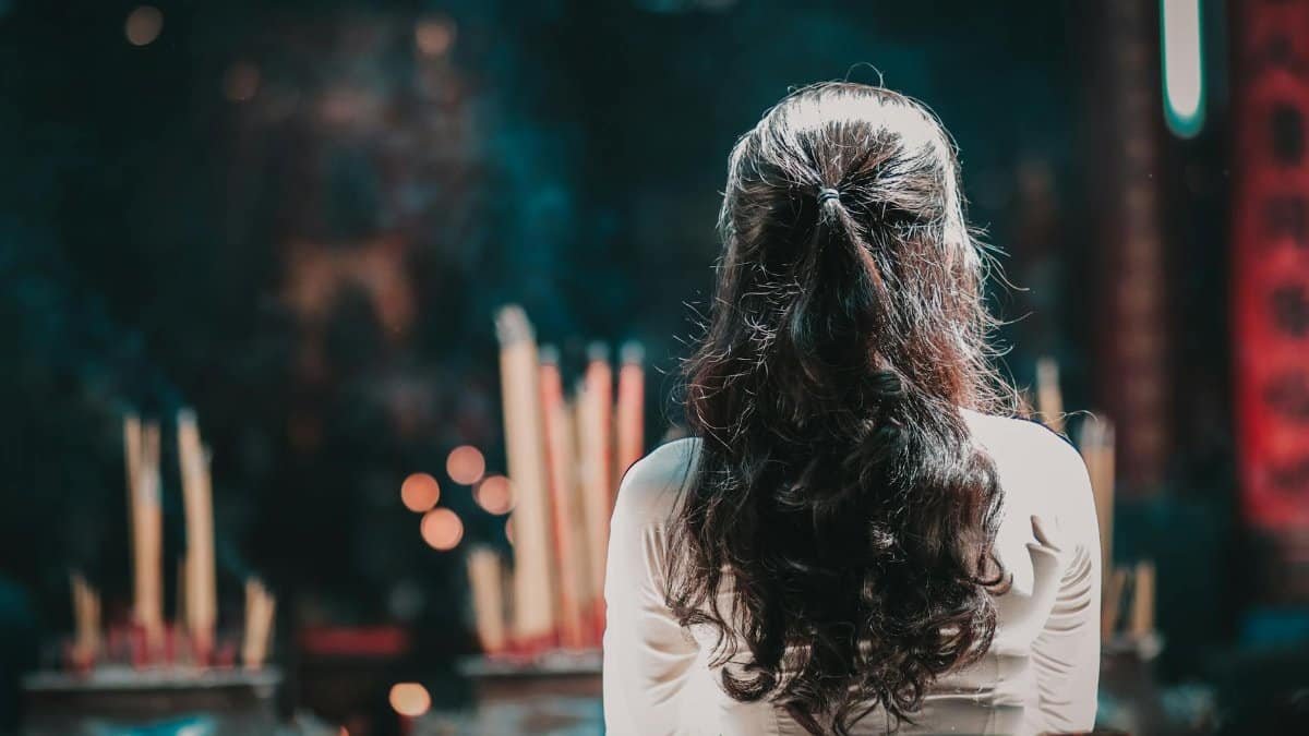 A woman with long hair is seen from behind praying inside a Vietnamese temple, surrounded by incense.