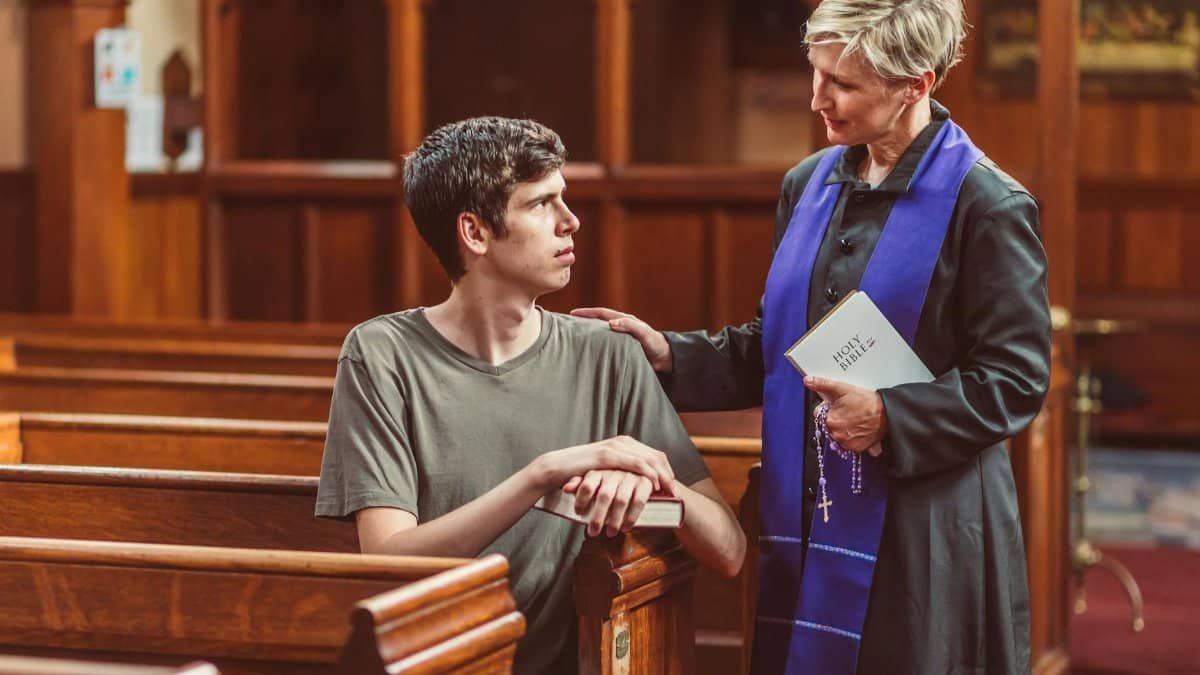 A clergy member comforts a young man in a church pew, discussing faith.