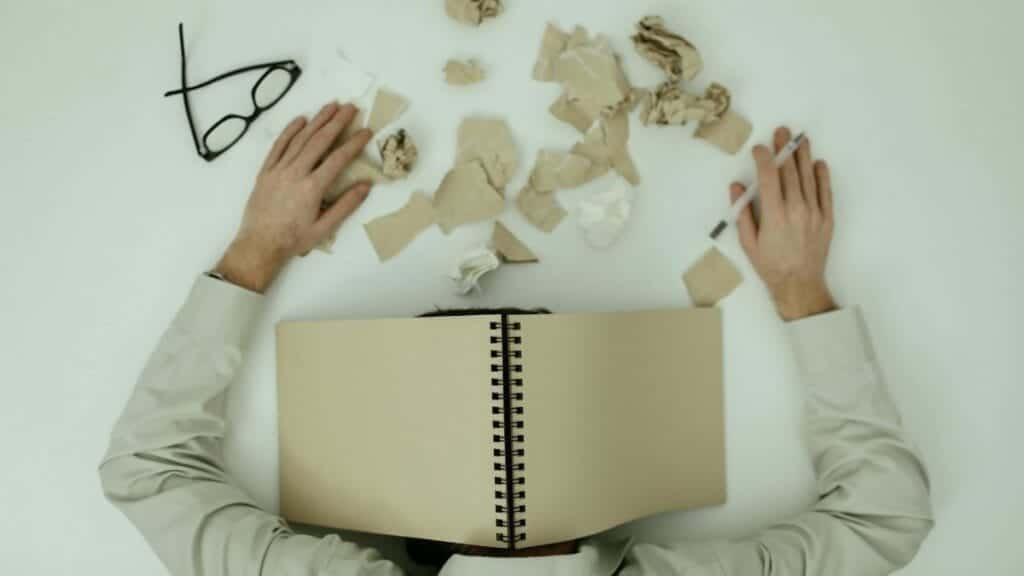 Overhead view of an exhausted man resting with a notepad over his head and crumpled papers around.