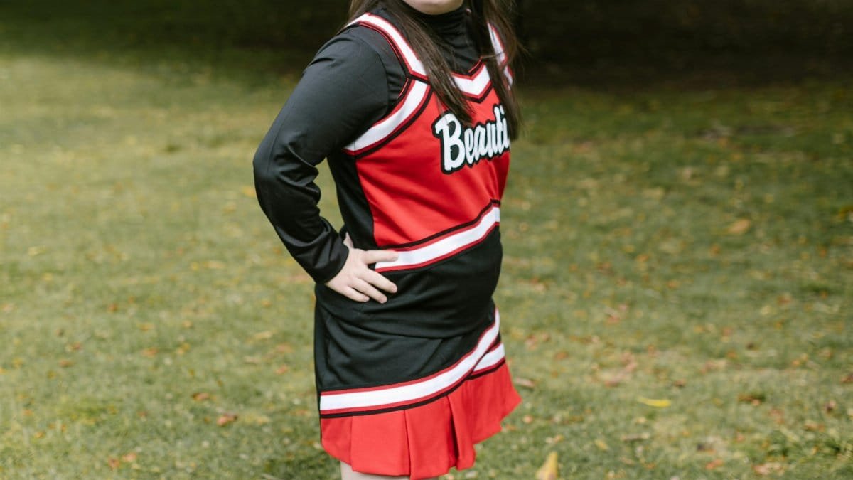 A cheerful young girl with Down Syndrome in a cheerleader outfit, posing outdoors in a park. via Pexels