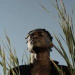 A serene portrait of a man meditating among tall grasses beneath a clear blue sky.