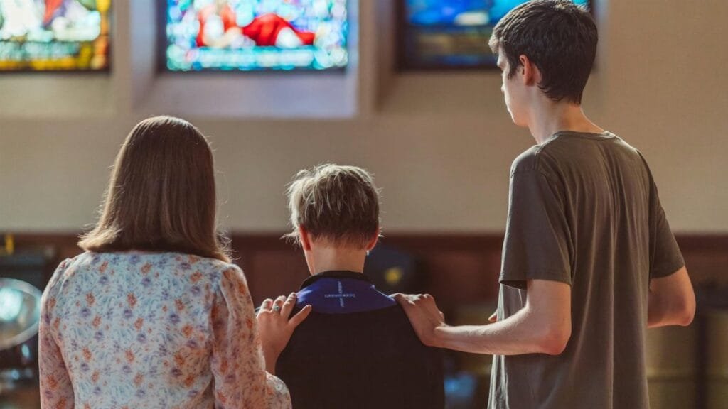 Three people gathered in prayer inside a church with vibrant stained glass windows.