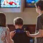 Three people gathered in prayer inside a church with vibrant stained glass windows.