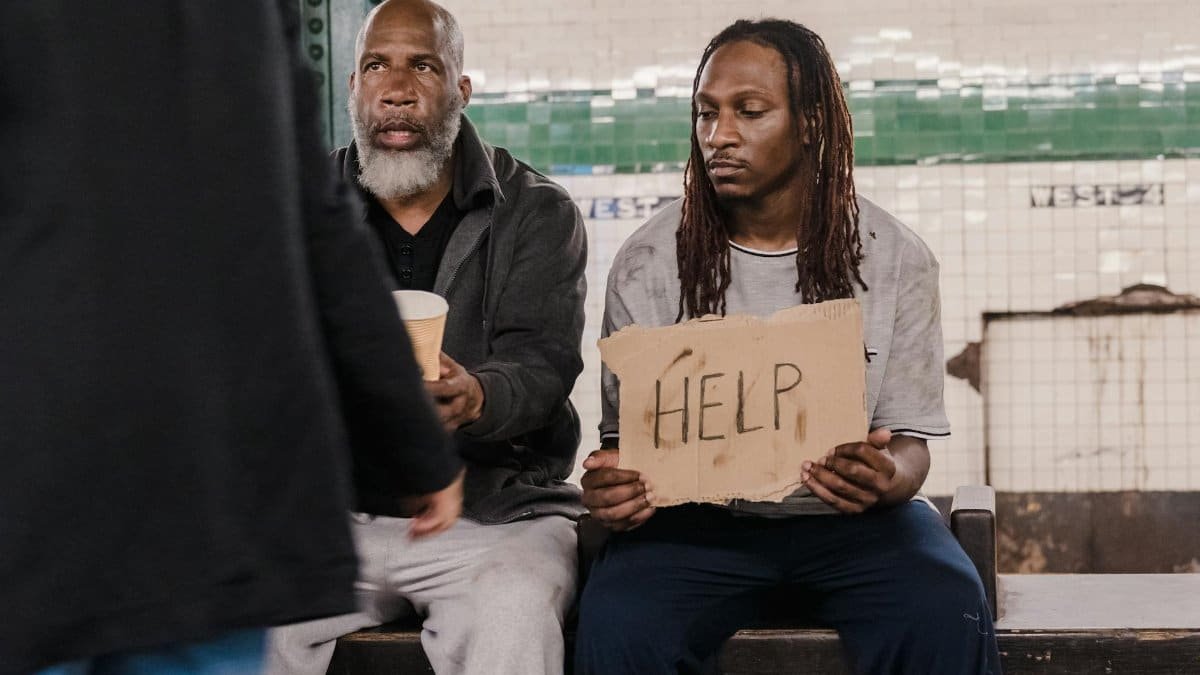Two men sitting on a bench holding a help sign, symbolizing social issues and homelessness.