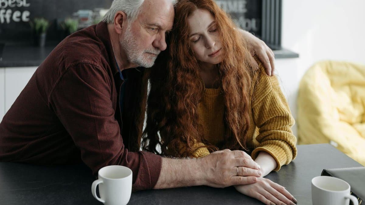 An elderly man consoles a young woman at a table in a warm, intimate indoor setting.